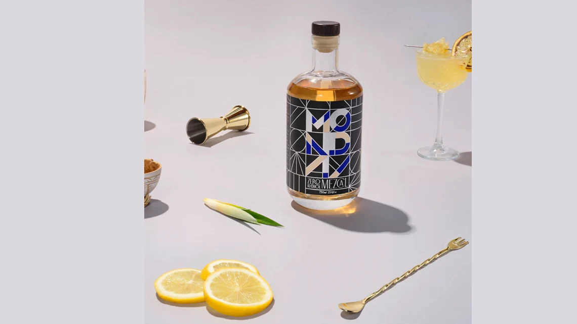 a bottle of Monday Zero Alcohol Mezcal surrounded by lemon slices, a cocktail, a mixer, and a spork