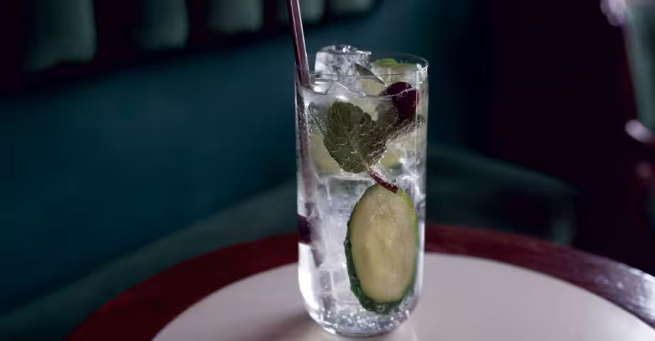 a glass of cuke & tonic with ice, cucumber slices, and basil leaves on a small round table