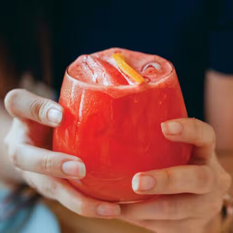 close-up shot of a woman’s hands holding a glass filled to the brim with a red-orange THC drink