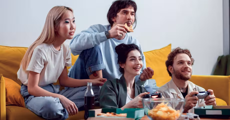 group of 4 friends seated around a yellow sofa playing video games and eating snacks