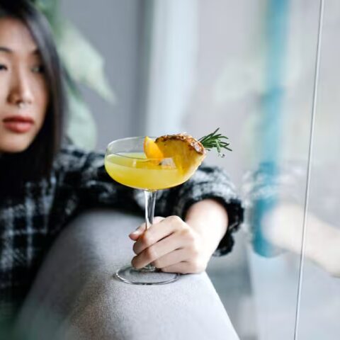 Asian woman leaning on a couch looking outside a window holding a pineapple mocktail