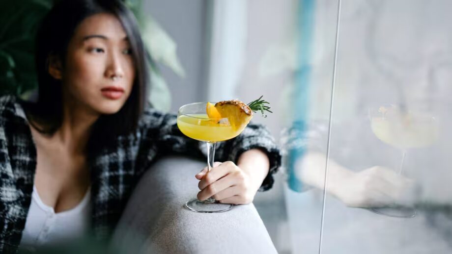 Asian woman leaning on a couch looking outside a window holding a pineapple mocktail